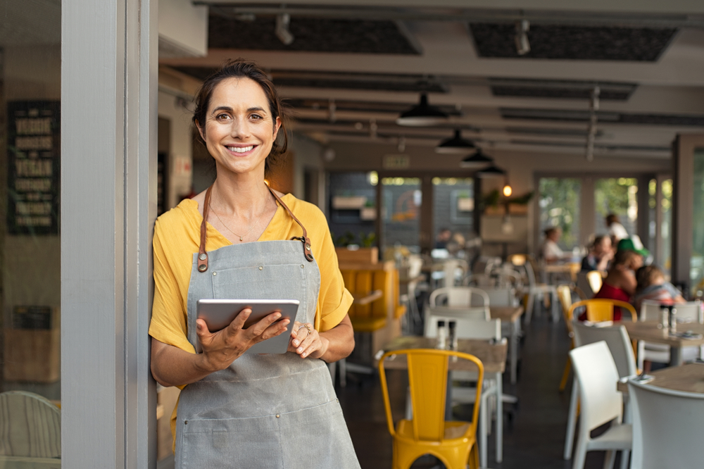 Portrait,Of,Happy,Woman,Standing,At,Doorway,Of,Her,Store. Employee Benefits Restaurant Dining Industry